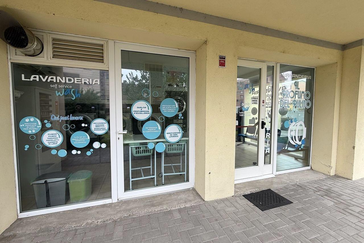 L'ingresso della lavanderia sotto il portico: le vetrine con le vetrofanie azzurre «Lavanderia self service wash», la porta a vetri e, a destra, l'entrata con la scritta «Il Profumo del Pulito»