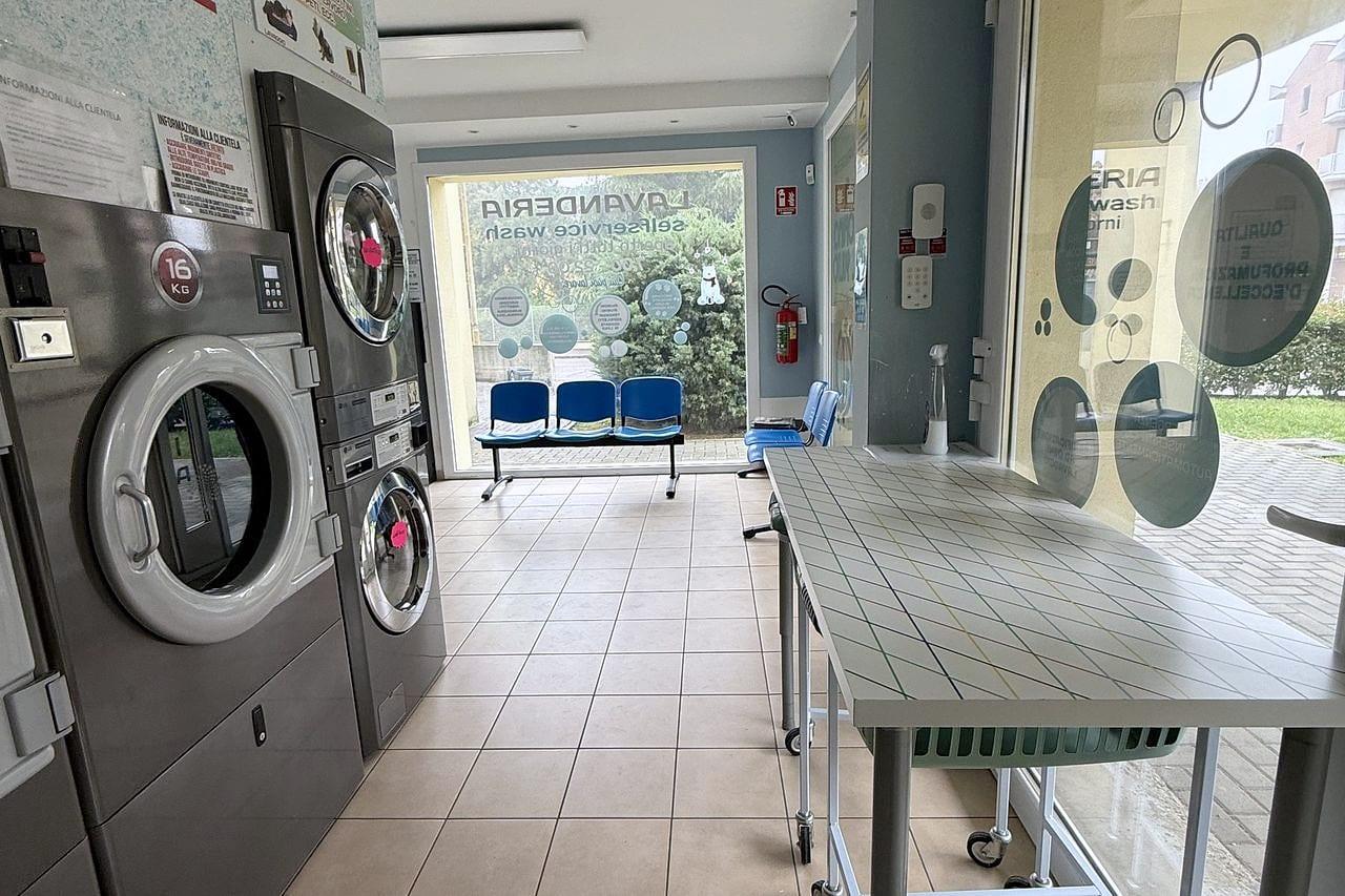 La zona d'attesa: in fondo le sedute blu davanti alla vetrina, a destra un lungo tavolo d'appoggio con i cesti, a sinistra le asciugatrici